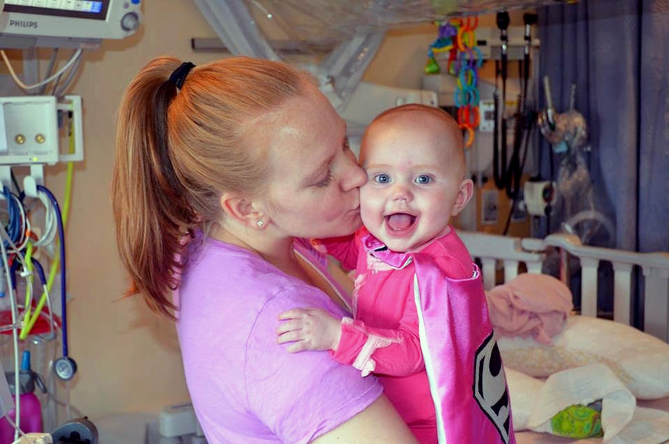 A woman, Kallie, with blond hair in a ponytail kisses the cheek of a smiling, blue-eyed baby, Sophie, wearing a pink superhero cape and facing the camera.