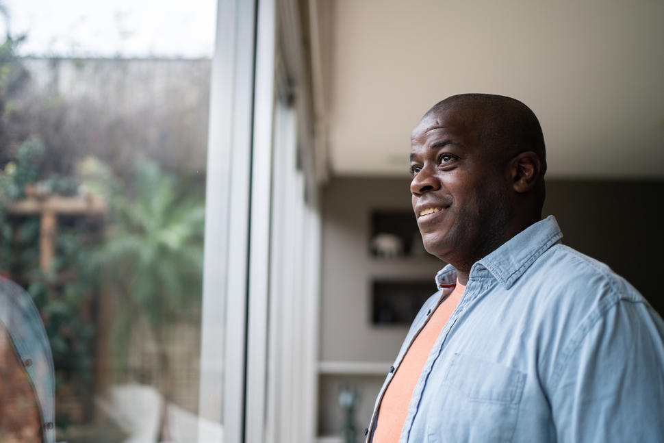 Man standing indoors by a window smiling and looking outside.