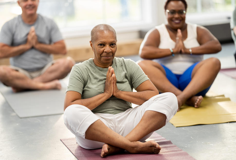 Woman with no hair and dark brown eyes sits cross-legged on a yoga mat with her hands in prayer position.