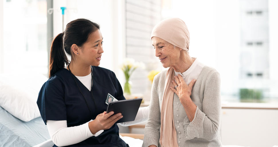 Enfermera con uniforme azul oscuro que señala una tableta electrónica mientras mira a una mujer con cáncer que sonríe con serenidad, tiene la mano izquierda sobre el pecho y lleva un pañuelo de color beige en la cabeza y viste ropa de color claro.