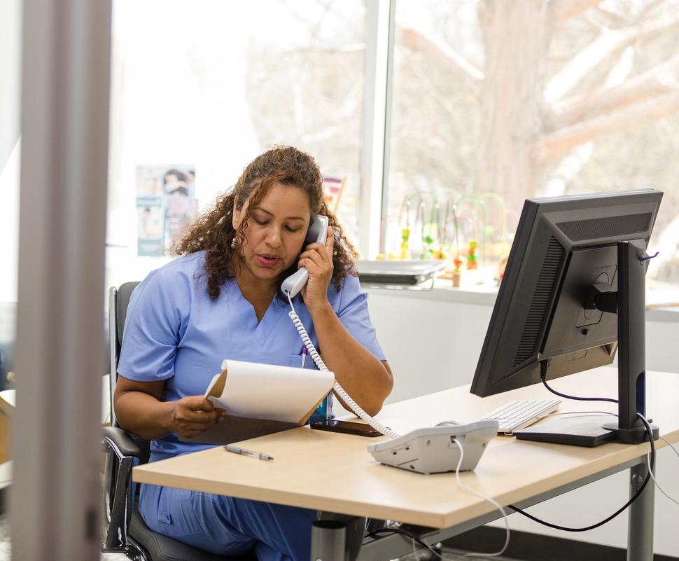 A woman in scrubs, sitting at a desk in a medical exam room, talks on a landline phone.