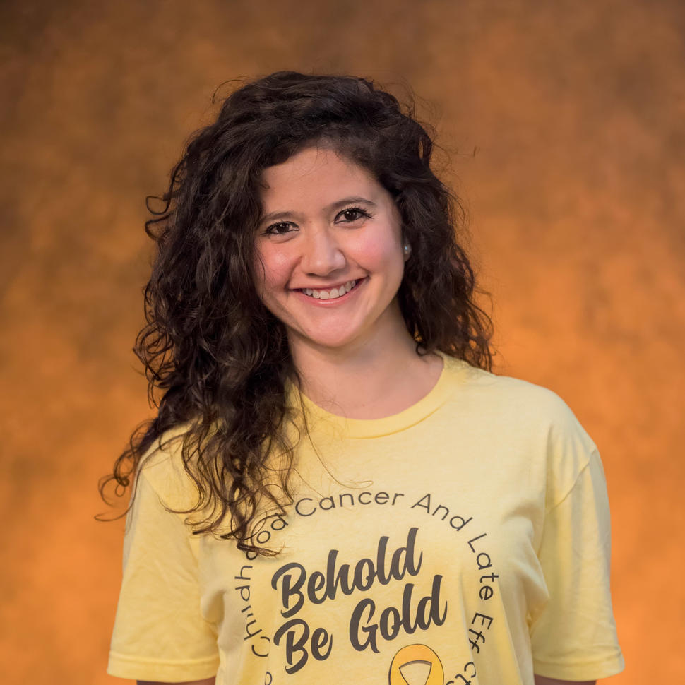 Juanita stands smiling at the camera, with dark brown, wavy hair and dark brown eyes, wearing a yellow T-shirt printed with “Behold Be Gold.”