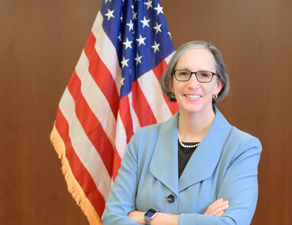 NCI Director Dr. Rathmell stands in front of the U.S. flag, smiling with her arms crossed
