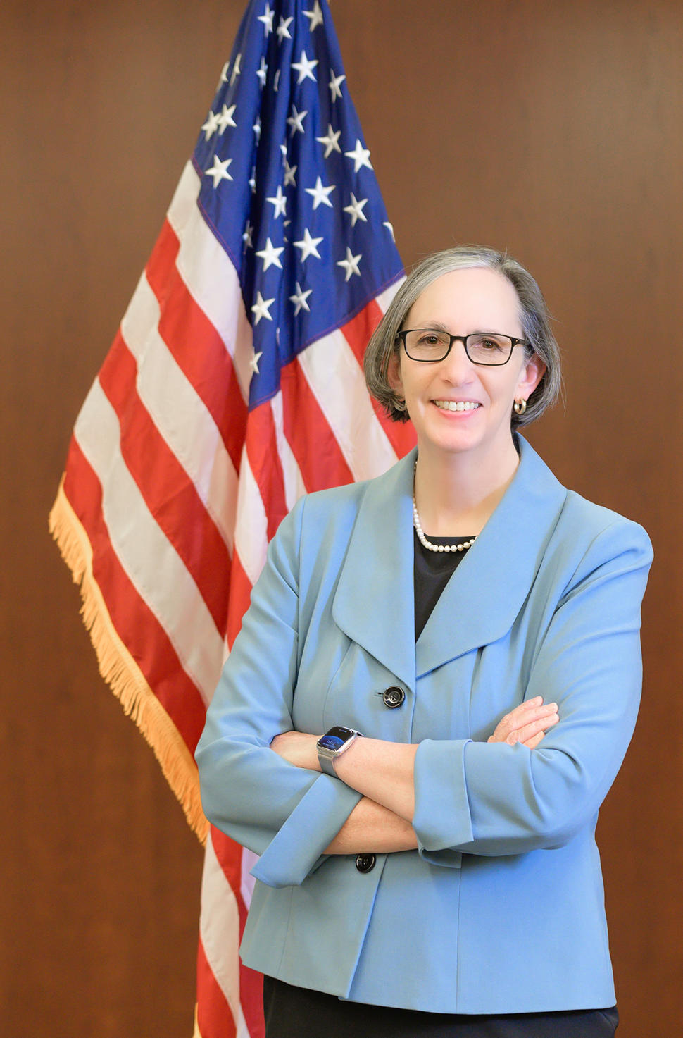 NCI Director Dr. Rathmell stands in front of the U.S. flag, smiling with her arms crossed