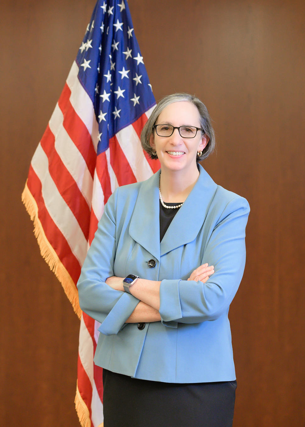 La directora del Instituto Nacional del Cáncer, la doctora Rathmell, se para frente a la bandera de los EE. UU., sonriendo con los brazos cruzados.