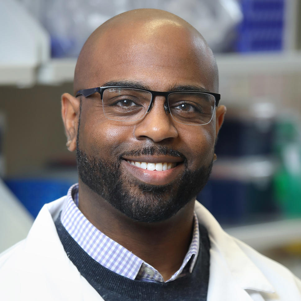 A photo of a man, Dr. Troy McEachron, who standing in a lab, smiling at the camera, and wearing glasses and a lab coat.