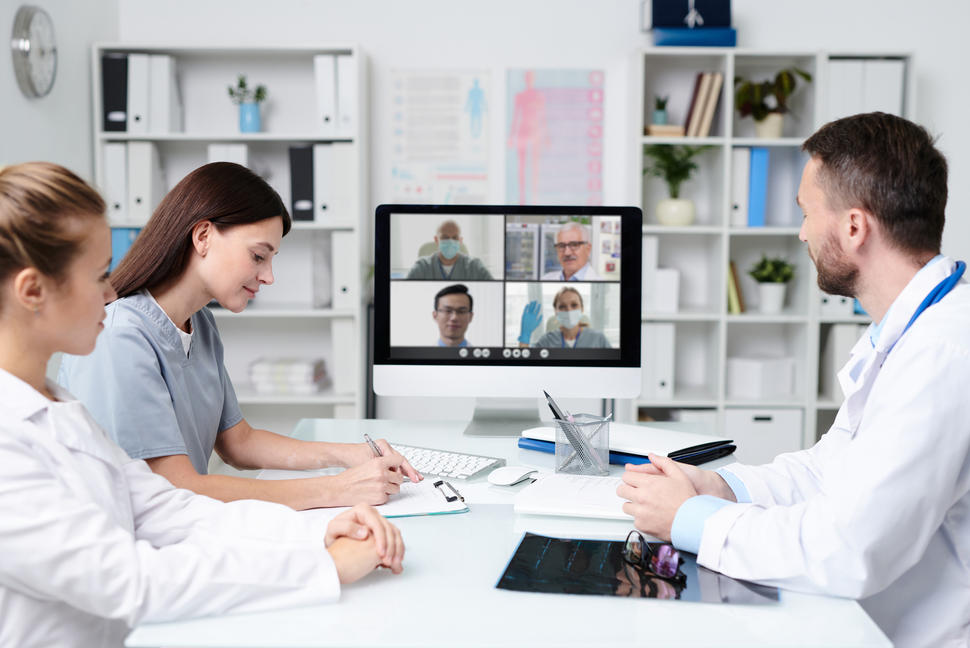 Three health care providers sit around a conference table having a videoconference with four health care providers working remotely. 