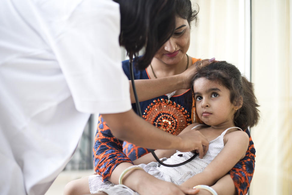 A health provider holds a stethoscope to a child's chest. The child sits on her mother's lap.