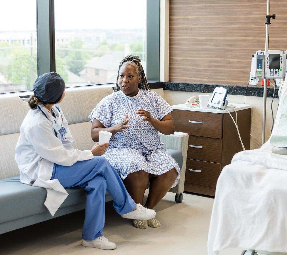 A middle-aged black woman in a hospital gown talking with a surgeon.