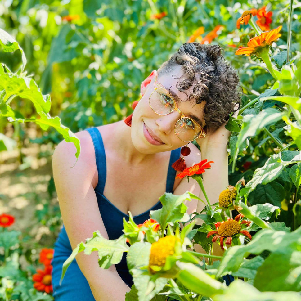 Hailey, with short, light-brown hair, stands in a field of sunflowers wearing a sleeveless blue top and yellow glasses and smiling at the camera.