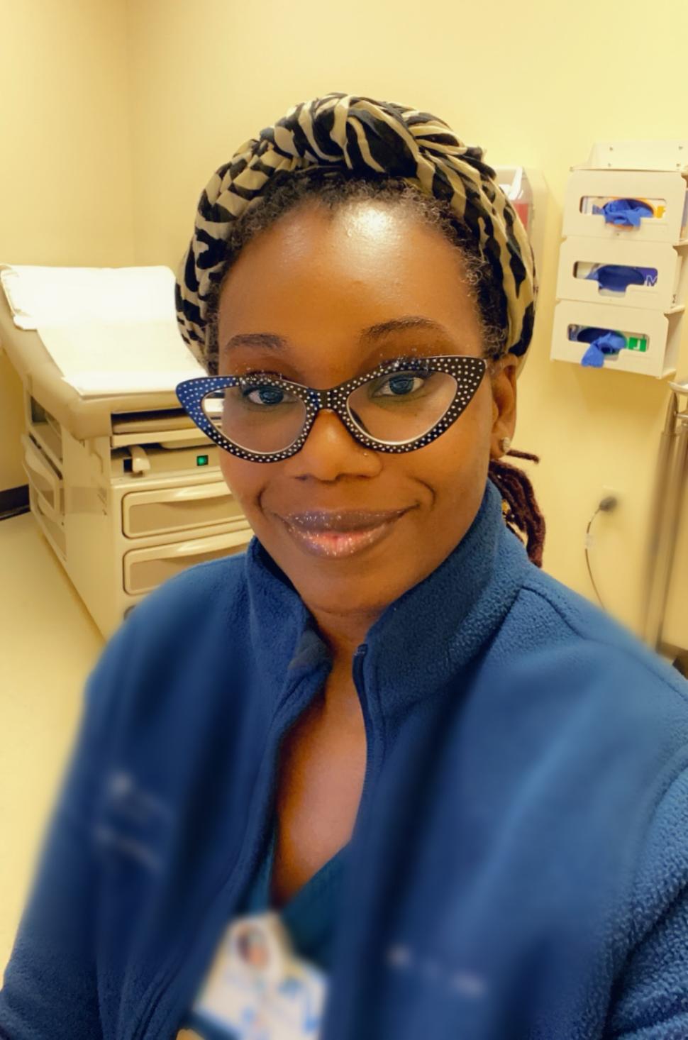 Photo of Asiyah Franklin. She is pictured in a medical exam room with an exam table behind her to the left and boxes of blue plastic gloves affixed to the wall behind her to the right.