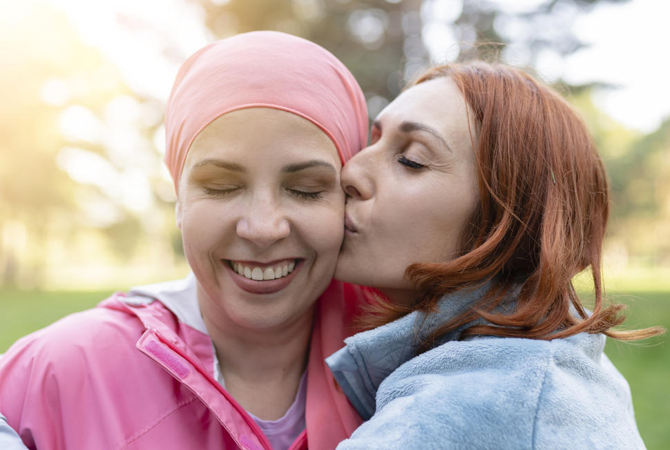 Photo of a smiling woman wearing a pink scarf on her head. Her eyes are closed and she is being embraced and kissed on the cheek by a woman with red hair.