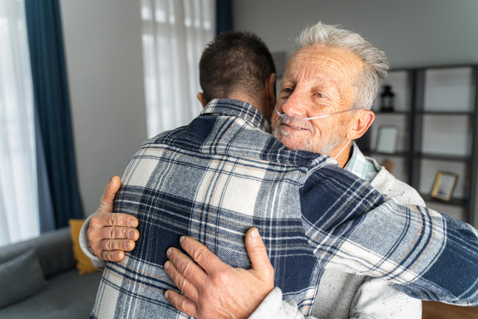 A frail older man with a tube in his nose peers over the shoulder of a younger man as they embrace.