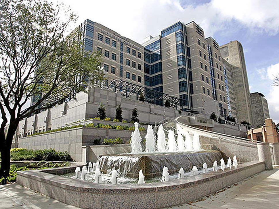 An exterior photo of the University of Texas MD Anderson Cancer Center. Fountains are in the foreground; in the background is the exterior of the building, which is mostly grey with some glass accents. Trees with some sparse leaves can be seen on either side of the building, and the sky is blue with puffy clouds.