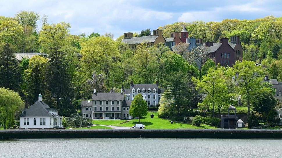 Cold Spring Harbor Laboratory Cancer Center buildings on a green hillside overlooking a waterfront, surrounded by dense trees under a partly cloudy sky.