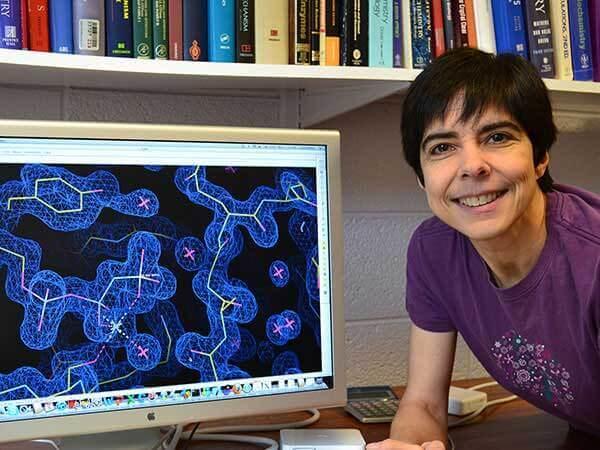 Carla Mattos looks at the camera and smiles. She has short black hair and wears a purple shirt. She’s next to a computer monitor with a complex visualization it. A bookcase full of books can be seen behind her.