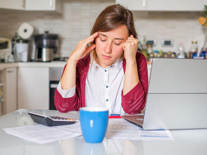 Young woman sitting at the kitchen table, looking down at several bills, a calculator, and a laptop.