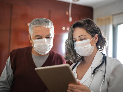 Male patient and female doctor wearing masks view a tablet together.