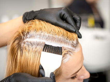 Hair color is applied to a woman's hair at the part line using a brush.