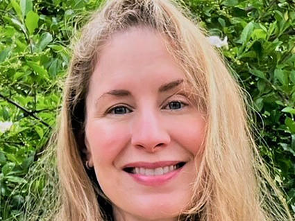 Woman with long blonde hair wearing a white blouse is smiling at the camera and stands in front of green foliage.