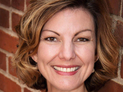 Woman with short, brown hair wearing a blue and white top smiling at the camera while leaning against a brick wall.