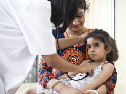 A health provider holds a stethoscope to a child's chest. The child sits on her mother's lap.
