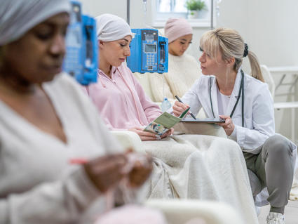 Three women sit in a clinic having IV infusions. A health provider kneels beside and chats with one young woman who is looking at a pamphlet.