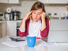 Young woman sitting at the kitchen table, looking down at several bills, a calculator, and a laptop.