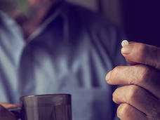A close up of a hand holding up a small, round, white pill. The hand and pill are in focus. In the background, out of focus, is another hand holding a cup and button-up shirt. Part of the man’s neck can be seen as well.