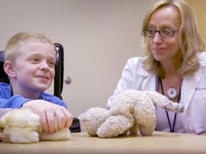 Brigitte Widemann (a light-skinned, blonde, female doctor in a white lab coat) is smiling and looking at a boy in a blue, long-sleeved shirt who is playing with two plush animals while talking to someone off camera.