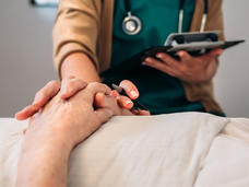 A nurse touches the hands of an elderly male patient laying in a hospital bed.