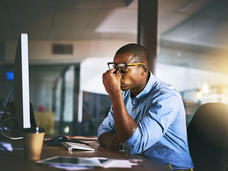 A man dressed in business casual clothing holds his hand against his face, pushing up his glasses and indicating that he is frustrated or stressed. He is sitting at his desk in front of his computer. The background indicates a business office.