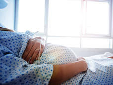 Foto de medio cuerpo de una mujer embarazada puesta una bata de hospital con una mano sobre su barriga, en la misma mano tiene un IV.