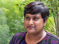 Deepa Prasad stands in front of a green, wooded background while looking at something off camera to her right. She is a medium-skinned woman with short, dark hair and is wearing a dark blue top with a light purple brushstroke print. 