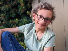 Micah is a young, light-skinned, non-binary person with short curly hair and glasses. They are wearing a mint green t-shirt and jeans and are smiling at the camera. 