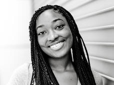 Black and white photo of young, Black woman with braids, standing against a building.