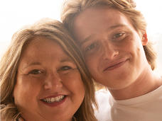 Lisa (a blonde, white woman) stands smiling and leaning into son, Jace (a blonde, white, young adult man).