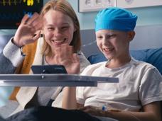 young person with cancer in hospital bed and health care provider looking at a tablet, smiling and waving