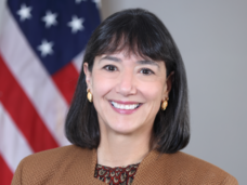 Woman (NCI Director Dr. Monica Bertagnolli) with dark hair wears a brown jacket over a patterned top and smiles at the camera.