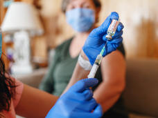 pair of gloved hands fills a syringe while a woman in a mask waits in the background