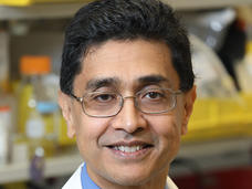 A man with medium brown skin tone and dark hair, wearing glasses, a blue shirt, a navy-blue tie, and a lab coat, is standing in a lab smiling at the camera.