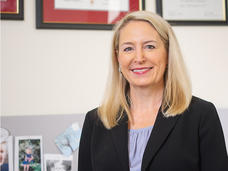 A professional headshot of Dr. Lynne Wagner, a White woman with a light skin tone and blonde hair, wearing a black blazer and blue top while smiling at the camera and standing in her office.