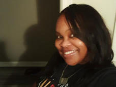 A black woman wearing a black t-shirt and chain necklace, taking a selfie in her home.