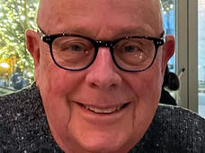 White man with glasses and grey knit sweater smiling at camera while sitting in front of glass doors. Outside are trees covered in white lights.