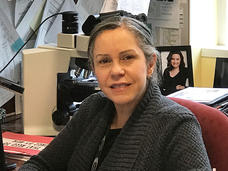 Puerto Rican woman (Nilsa Ramirez) sits at a desk with a microscope, framed photo, and papers tacked to a bulletin board behind her.