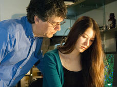 A young woman sits looking and pointing a pen at a paper on a desk in a lab office. Her father leans over her shoulder looking at the same paper. 