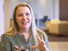 A woman (Dr. Emily Tonorezos) with long blond hair wears a muted green button-up blouse and sits in a chair, smiling and talking with her hands.