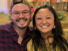 Woman (Sarah) with long black hair, earrings, and peach-colored blouse with blue and white flowers sits next to her husband. Both smile at the camera.