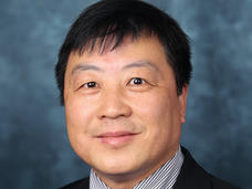 A man (Dr. Xiao-Nan Li) with dark hair and brown eyes wears a striped collared shirt, tie, and dark jacket and smiles at the camera.