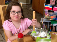 A girl, Abby, wearing glasses and a pink T-shirt, stirs ingredients in a metal bowl with a rubber spatula and smiles at the camera.